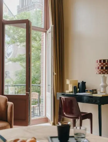Bright hotel room with open balcony doors, natural light, and a small desk beside the window.